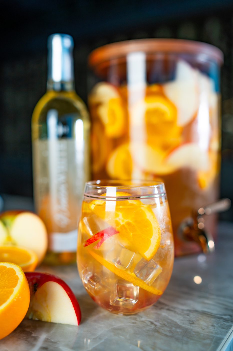 Glass of iced fruit‑infused white wine with orange and apple slices, pitcher dispenser and wine bottle blurred behind.