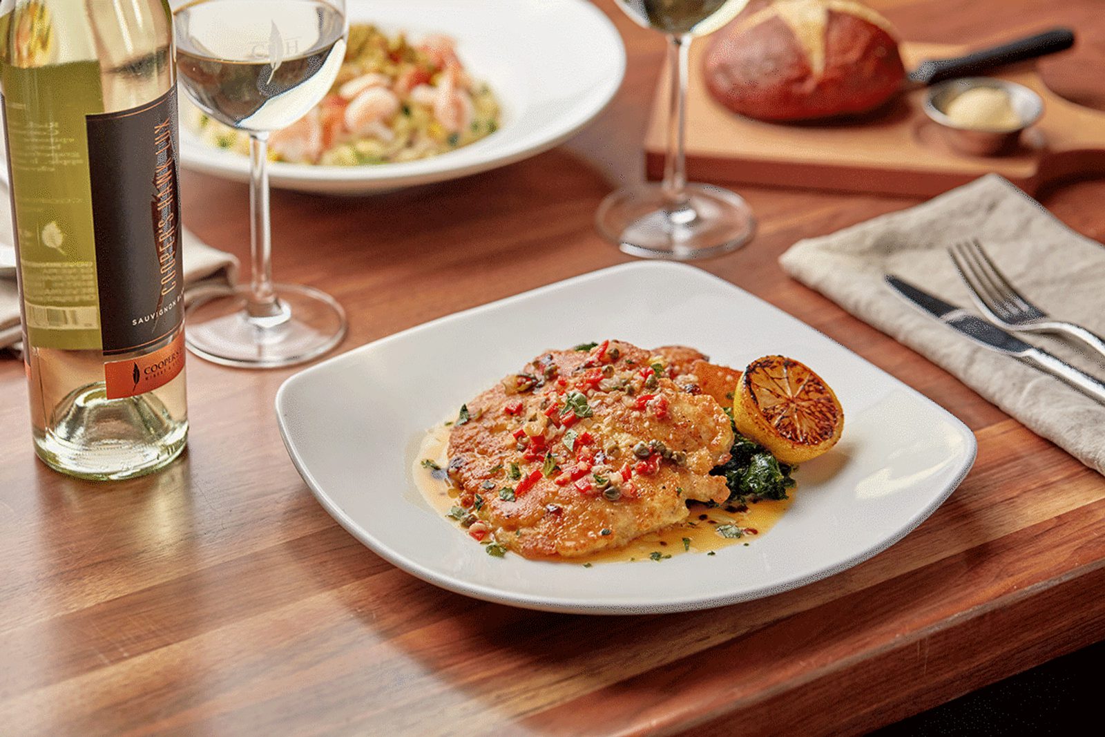 Plated grilled fish with herb sauce and charred lemon on a wooden table, with white wine glasses, a bottle, and a folded napkin with cutlery.