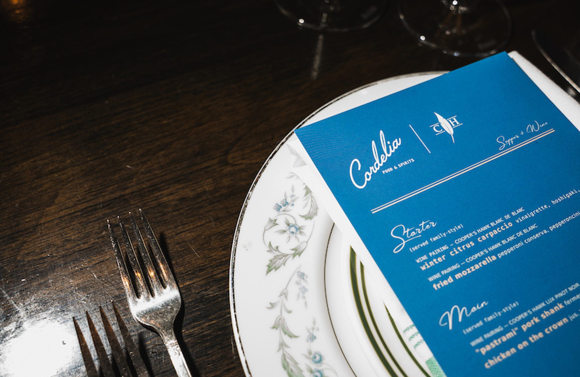 Blue menu titled 'Cordelia' on floral-rim china plate, two forks to left and two wine glasses above on dark wood table